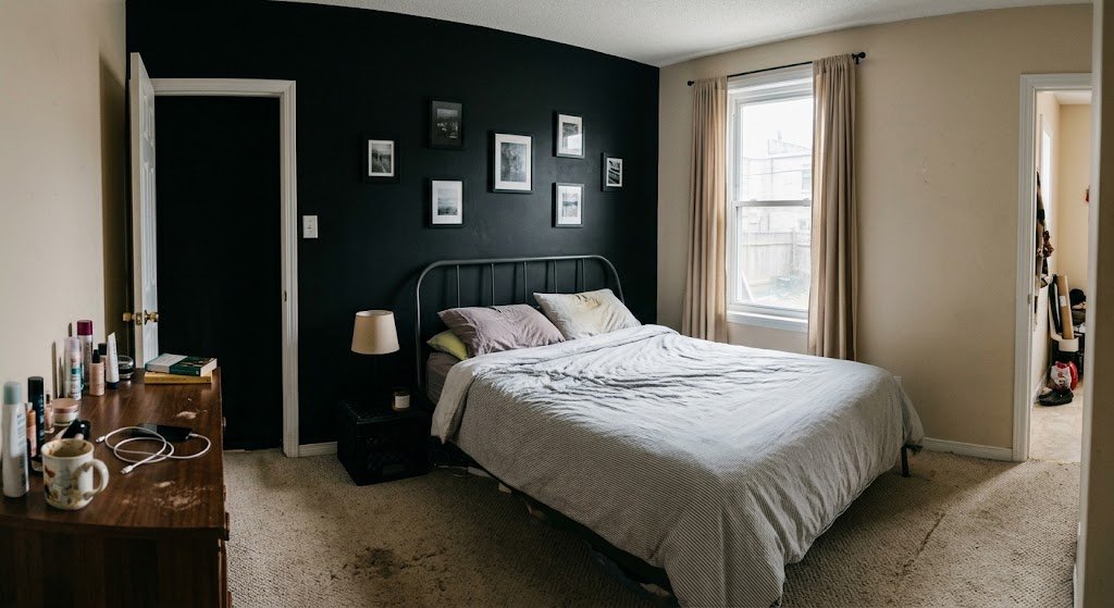 A dimly lit bedroom featuring an unmade bed set against a black accent wall with small, disjointed picture frames.