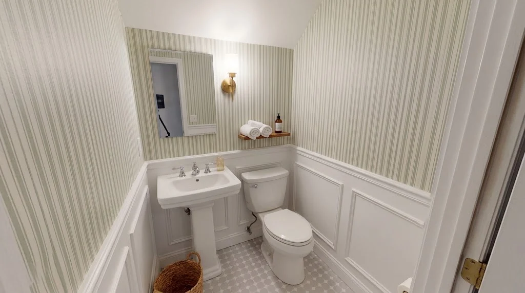 A naturally lit view inside a windowless small half-bath with vertical sage stripe wallpaper.